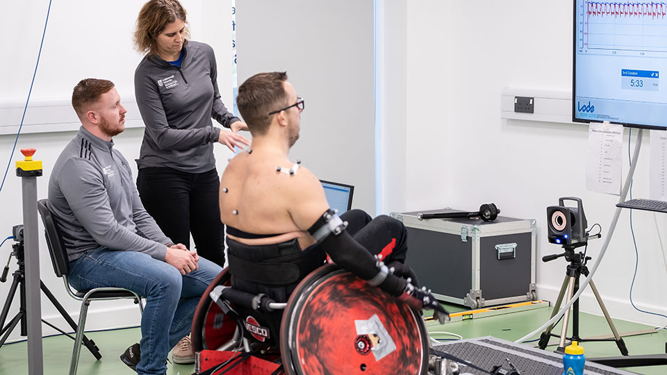 A man in a wheelchair on an adapted treadmill being monitored by two researchers from the Peter Harrison Centre for disability sport