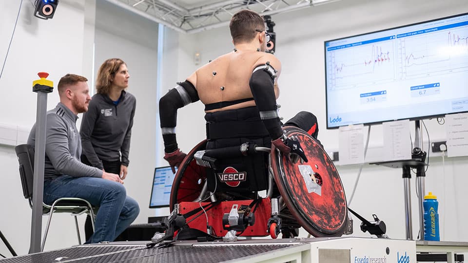 A para athlete in a wheelchair on a treadmill with two academics looking at data on a big screen