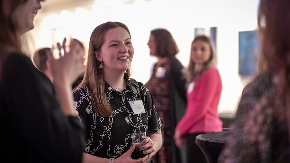 An alumna is in the centre of the image smiling. Others are looking towards her and more people are in the background.