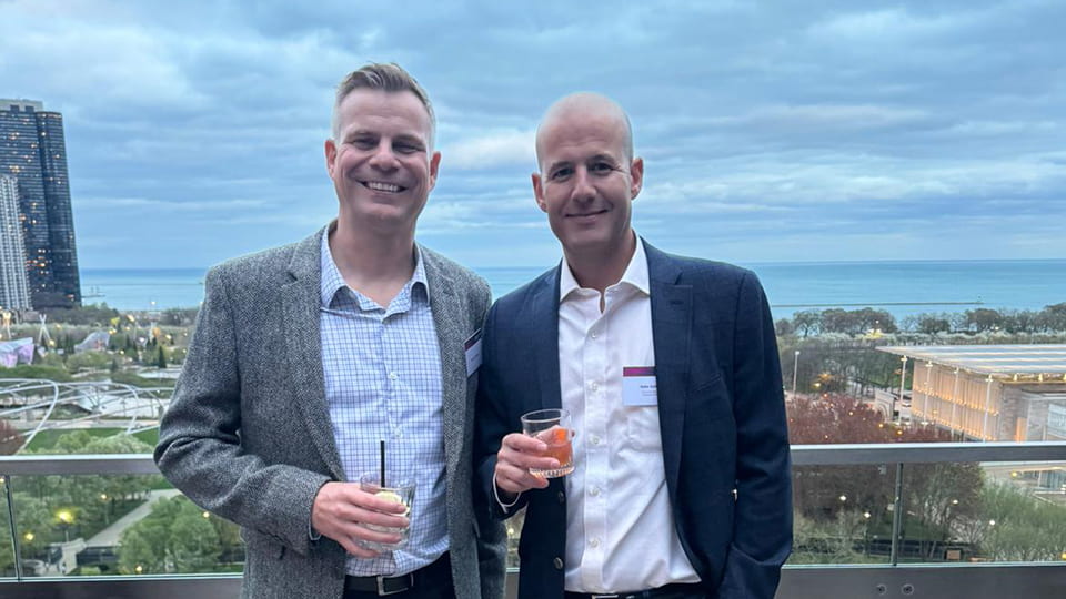 Two people standing together smiling holding drinks. There is a view of a cityscape in the background.