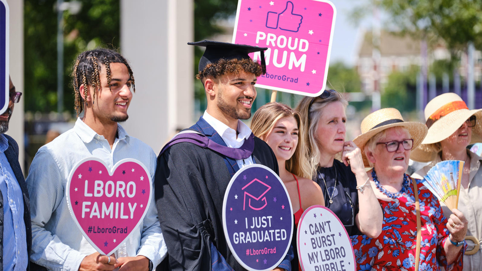 A group of people standing and smiling holding LboroGrad signs at graduation in summer 2022