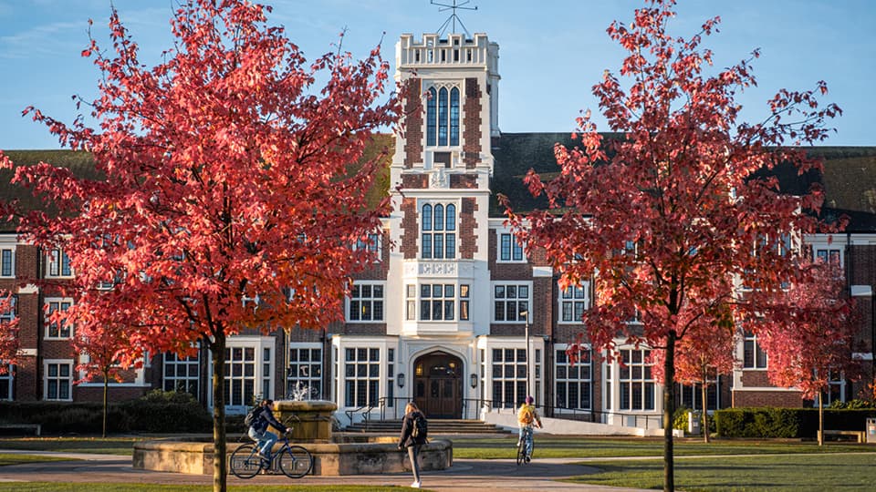 The Hazlerigg Building with two trees in front that have red autumnal leaves on. People walk and cycle on the paths in front.