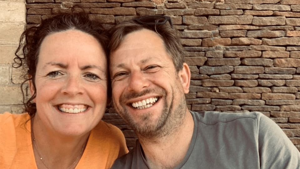 A close up of Carmen and James grinning at the camera. They are both sat against a brick wall as a background.