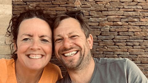 A close up of Carmen and James grinning at the camera. They are both sat against a brick wall as a background.