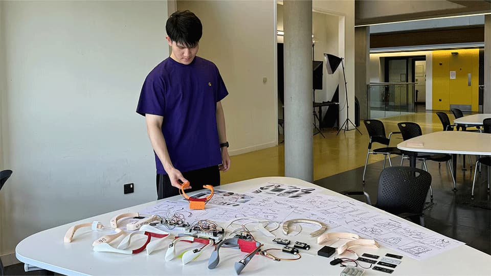Zuming is wearing a navy t-shirt and is standing at a table with prototypes of his project SALANCE, an uneven shoulder detection device.