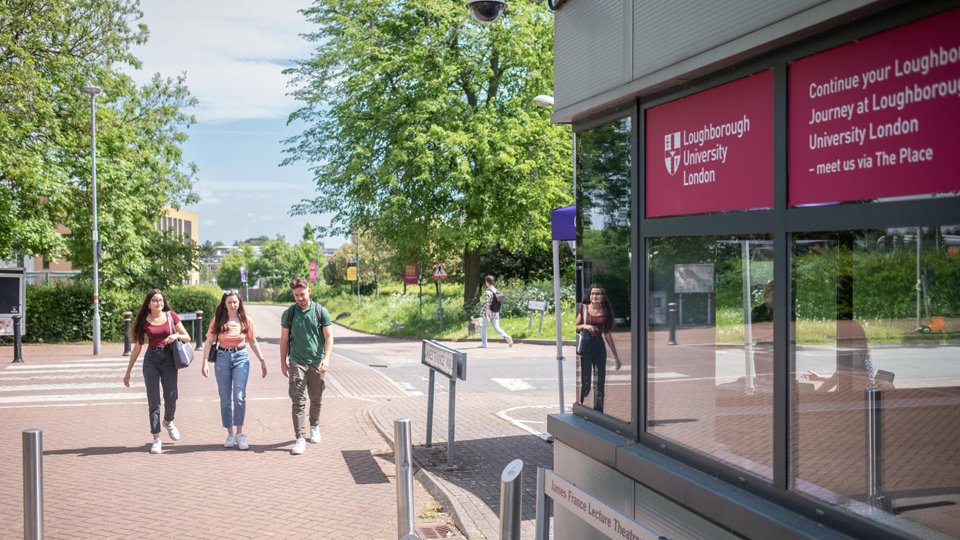 Three students walking on campus. There is a building, a road and trees.