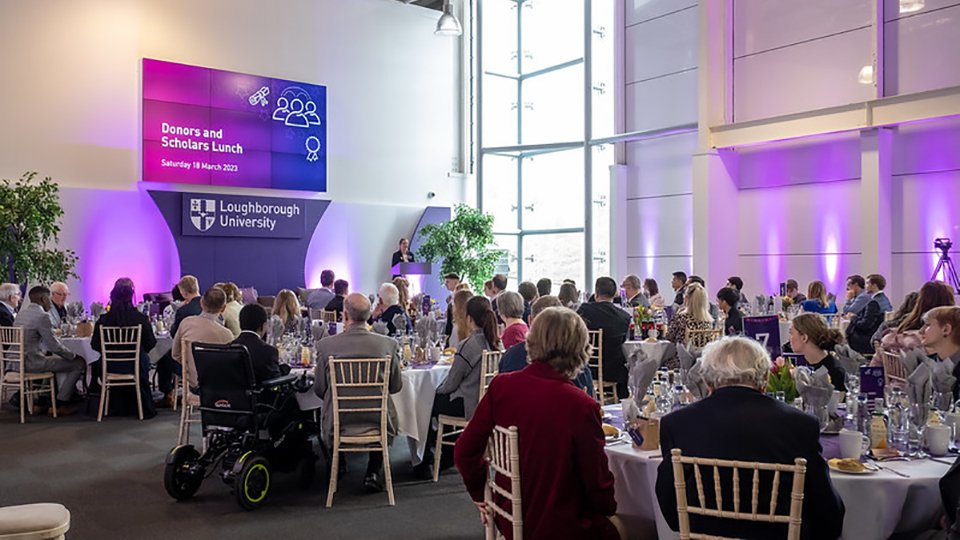 Groups of people seated at circular tables facing towards a stage where someone stands to address the room.