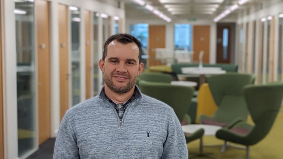 Adriano standing up looking at the camera. He is in focus and in the background is a room with tables and chairs which are a blurry in the background.