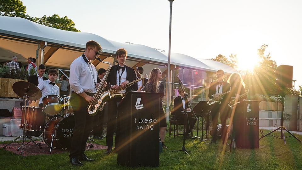 Student jazz band Tuxedo Swing. The people have a range of instruments are outdoors on grass.