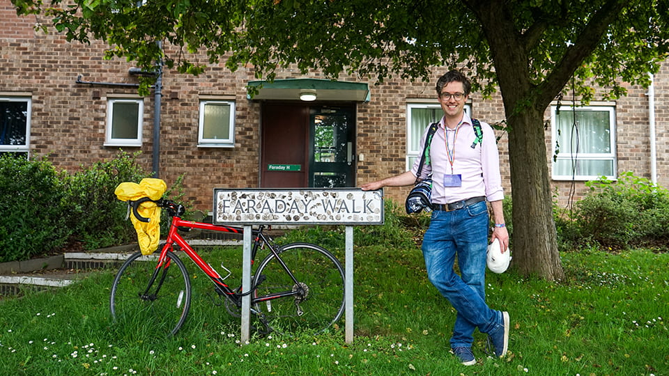 A person stands next to a street sign that reads Faraday Walk. There is a bike leaning on the sign and a building behind.