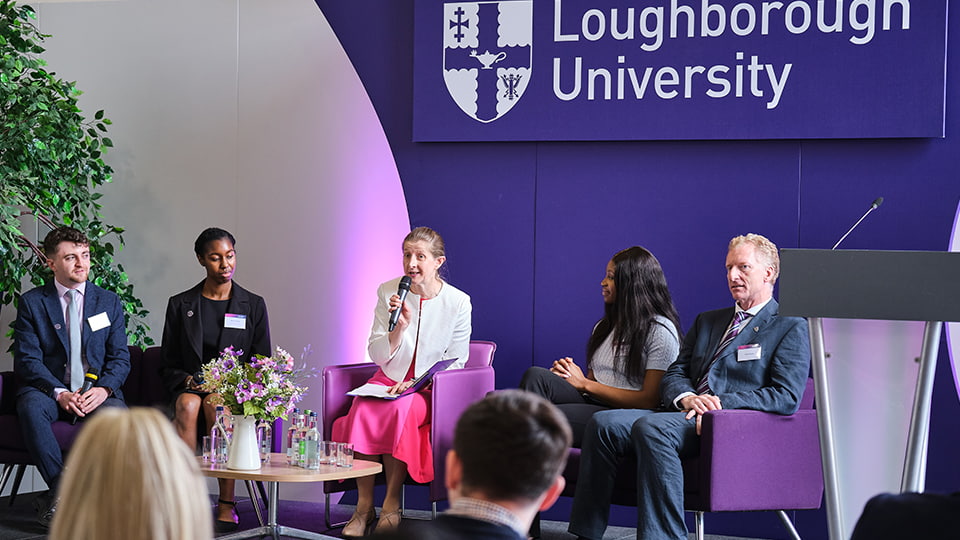 Five people seated on a stage. There is a large Loughborough University logo on the wall behind them.
