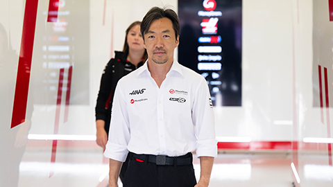 Ayao Komatsu. There is a person walking behind him and a screen on the wall.