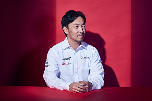Ayao Komatsu with his hands placed together on a table. The backdrop and table are red. He looks to the side.