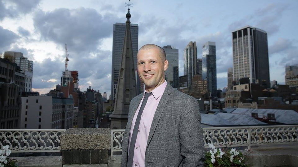 Pete Fleming stands in front of a cityscape. He wears a suit.