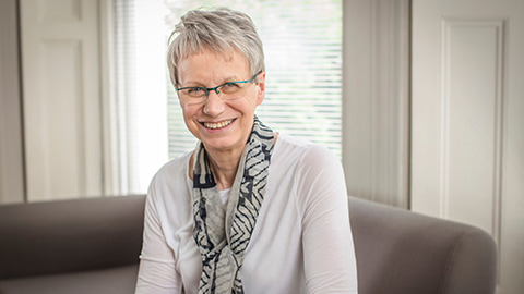 Professor Dame Judith Petts smiling, seated on a sofa with a window behind. She wears a scarf and spectacles.