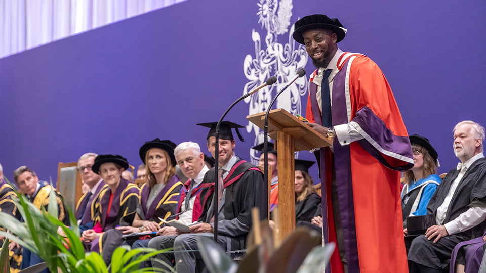 Nana Badu wearing a red gown standing at a podium. There are people seated behind him also wearing gowns.