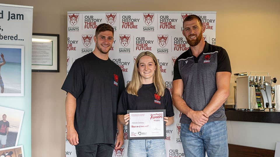 Heledd is standing with James Grayson (Ex-Northampton Saints player) and Tom Wood (Ex-Northampton Saints and England player) from Northampton Saints Foundation. The three of them are smiling in front of a banner that says 'Northampton Saints Foundation. Our history their future.'