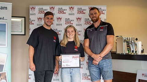 Heledd is standing with James Grayson (Ex-Northampton Saints player) and Tom Wood (Ex-Northampton Saints and England player)  from Northampton Saints Foundation. The three of them are smiling in front of a banner that says 'Northampton Saints Foundation. Our history their future.'