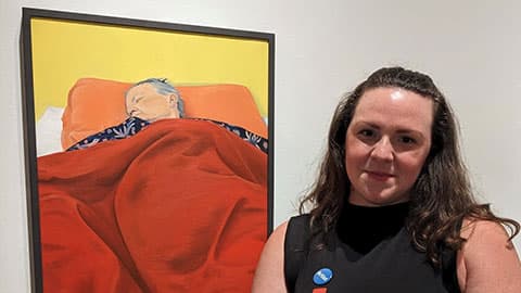 Laura is at The National Portrait Gallery in London standing in front of her painting called ‘I’ll Never Not Miss You’. The painting is in a black frame and the painting is of her mum asleep in bed with a red blanket over her.