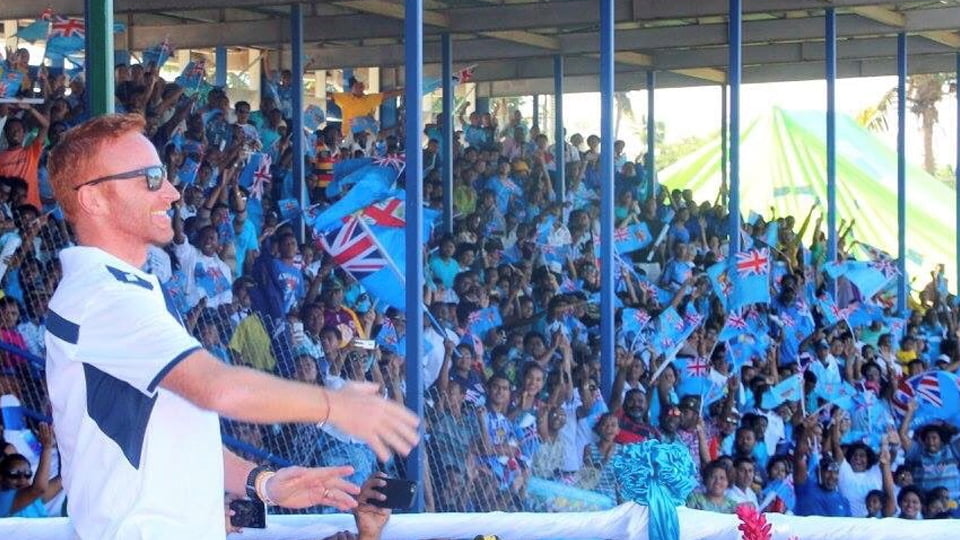 Ben Ryan wearing sunglasses and smiling. In the background there are many fans wearing blue tops and waving flags. They are in a stadium.