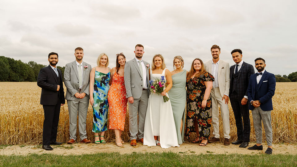 Danny is wearing a grey suit and Sian is wearing a white wedding dress. They are standing with friends and family on a patch of green grass.