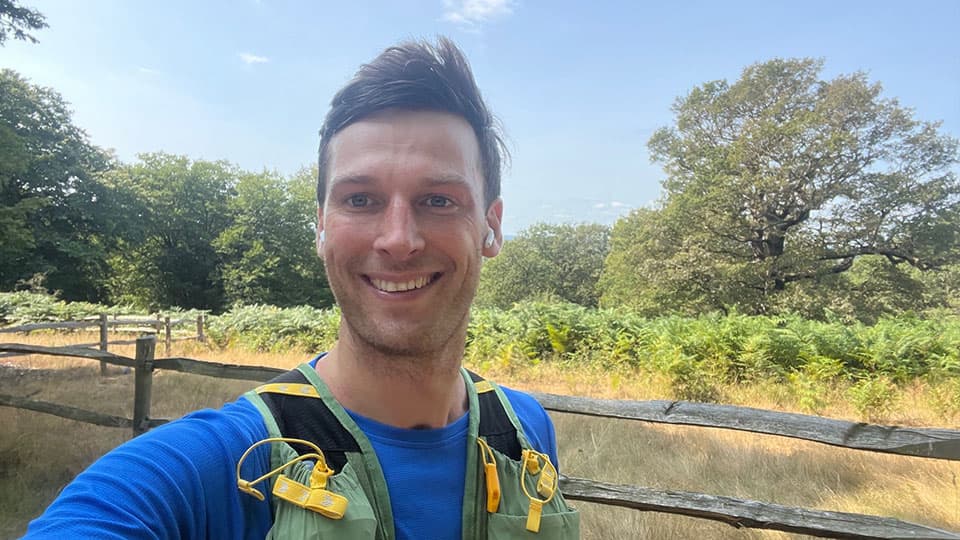 James is smiling at the camera and is running in a running vest. There is blue sky and he is surrounded by trees. The sun is out.