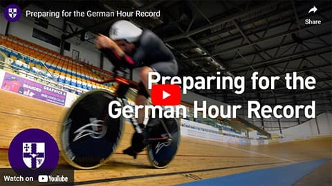 A screenshot of a YouTube video. There is a photo of someone riding on the track on a bike with text overlaying that says 'Preparing for the German Hour Record'