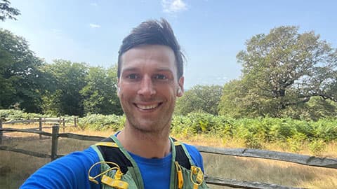 James is smiling at the camera and is running in a running vest. There is blue sky and he is surrounded by trees. The sun is out.