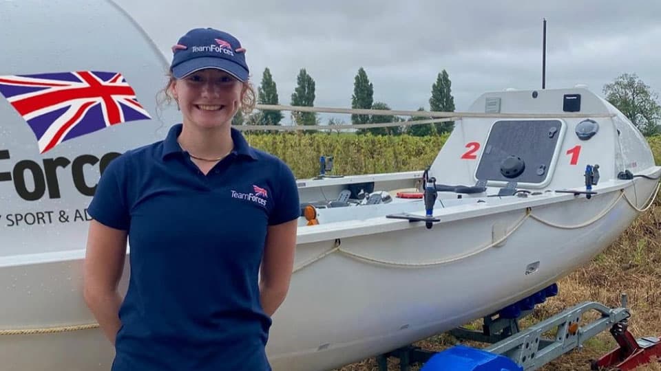 Zara is smiling and standing in front of a boat. She is wearing a navy hat and t-shirt with the Team Forces logo on them.
