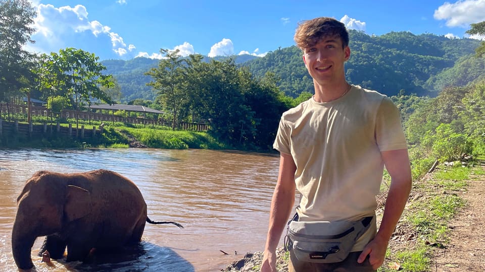 Jamie stands on the right with an elephant in water to the left. There is a beautiful scenery featuring water, trees, and blue sky.