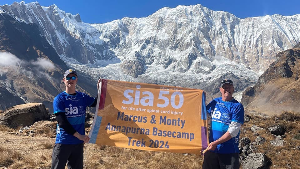 Marcus and his son standing in front of the Annapurna Base Camp with a banner which says 'sia 50 for life after spinal cord injury. Marcus & Monty Annapurna Base Camp Trek 2024'