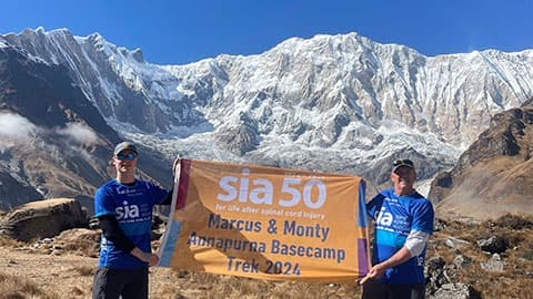 Marcus and his son standing in front of the Annapurna Base Camp with a banner which says 'sia 50 for life after spinal cord injury. Marcus & Monty Annapurna Base Camp Trek 2024'