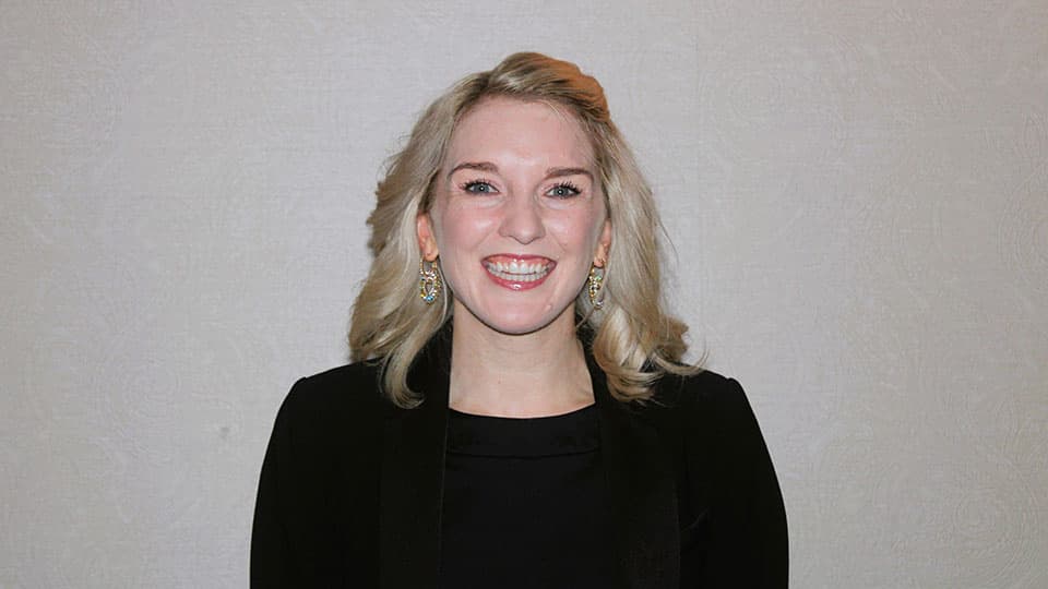 A headshot of Sarah Black-Smith. She is wearing black blazer and long earrings. She is smiling at the camera.