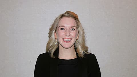 A headshot of Sarah Black-Smith. She is wearing black blazer and long earrings. She is smiling at the camera.