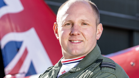 Flt Lt Rob Thomas wearing his green RAF Aerobatic Team suit. He stands in front of a red Arrows plane with his arms folded.