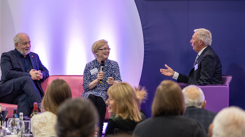 Barbara Slater seated on stage with Professor Nick Jennings and David Moorcroft. Barbara holds a microphone and looks towards David who is asking her a question.