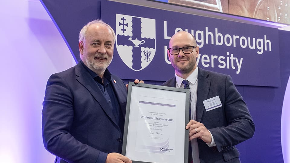 Professor Nick Jennings hands George Drake a certificate for Dr Herbert Schofield. They stand together in front of a Loughborough University banner.