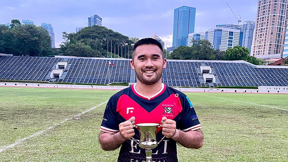 Callum holding the Presidents Cup on a rugby pitch. He is grinning and standing their in his rugby kit.
