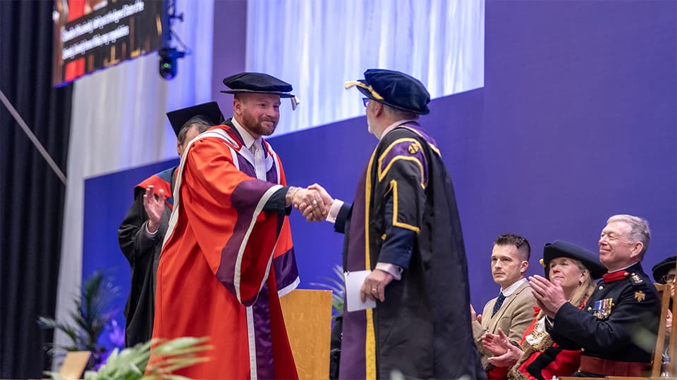Adam Peaty wearing a red gown and graduation cap shaking hands with the Vice-Chancellor on stage