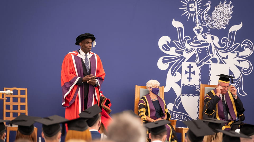 Professor Ivan Browne OBE stands on graduation stage wearing a red gown. There are people in face masks seated behind.