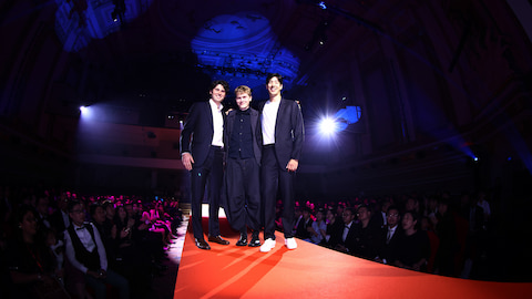 Three people stand on a red carpet. Either side are people seated. The room is dark.