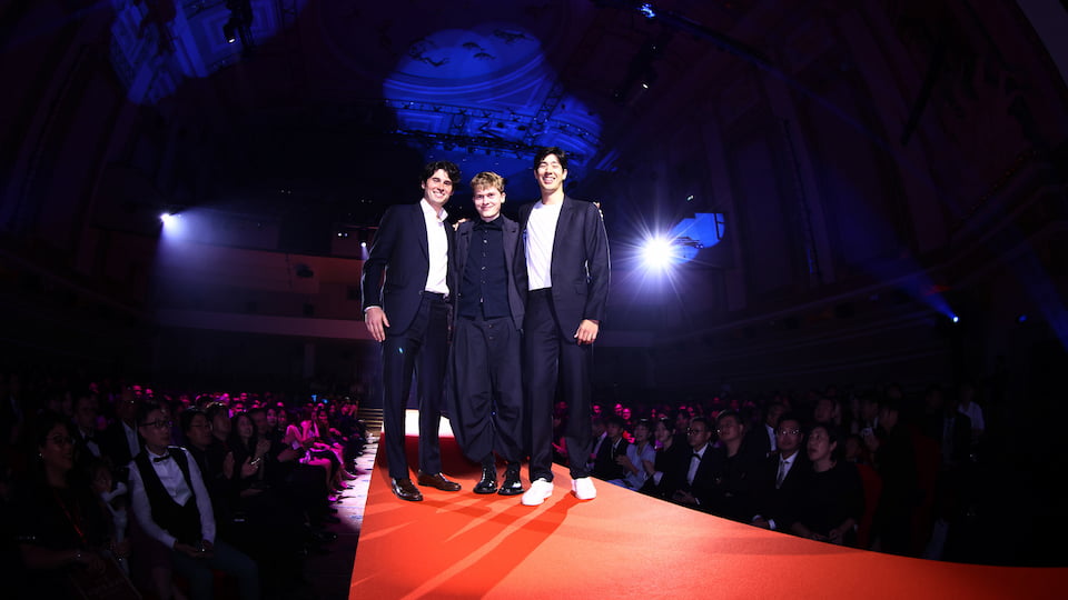Three people stand on a red carpet. Either side are people seated. The room is dark.