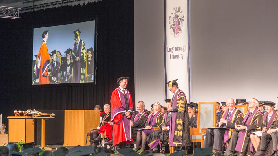 Professor Paul Brickell wearing a red gown standing on stage to receive his Honorary Degree. Lord Coe stands, too. People are seated behind them.
