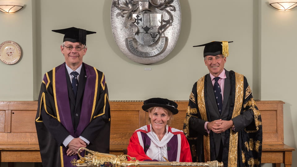 Penny Briscoe CBE seated at a table smiling wearing a red gown. Prof Chris Linton is on the left and Lord Sebastian Coe on the right, both wearing graduation gowns. A mace is on the table.