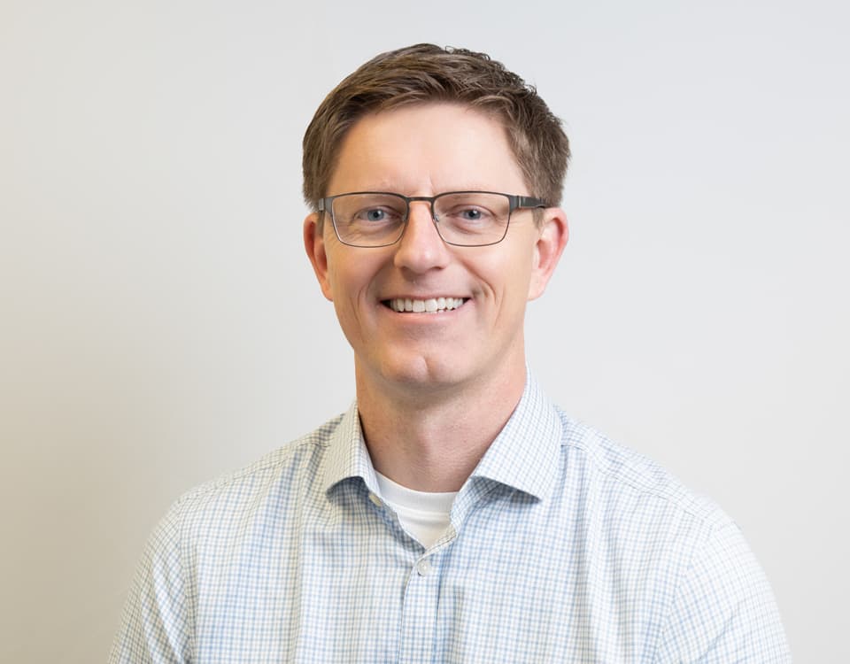 Jay Wakenshaw smiling, wearing a shirt. He is against a plain background. He wears glasses.