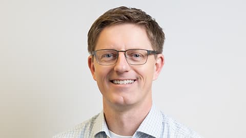 Jay Wakenshaw smiling, wearing a shirt. He is against a plain background. He wears glasses.