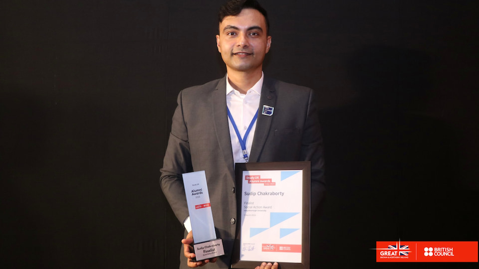 Sudip stands holding a trophy and certificate. He wears a suit. The background is black. There is a British Council logo in the bottom right corner.