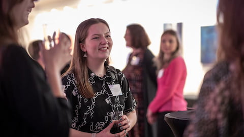An alumna is in the centre of the image smiling. Others are looking towards her and more people are in the background.