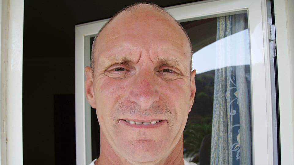 A close up of Bob Chappell BEM wearing a white t-shirt and smiling at the camera. He is standing outside in the sunshine.