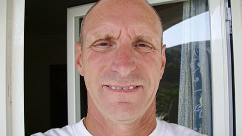 A close up of Bob Chappell BEM wearing a white t-shirt and smiling at the camera. He is standing outside in the sunshine.